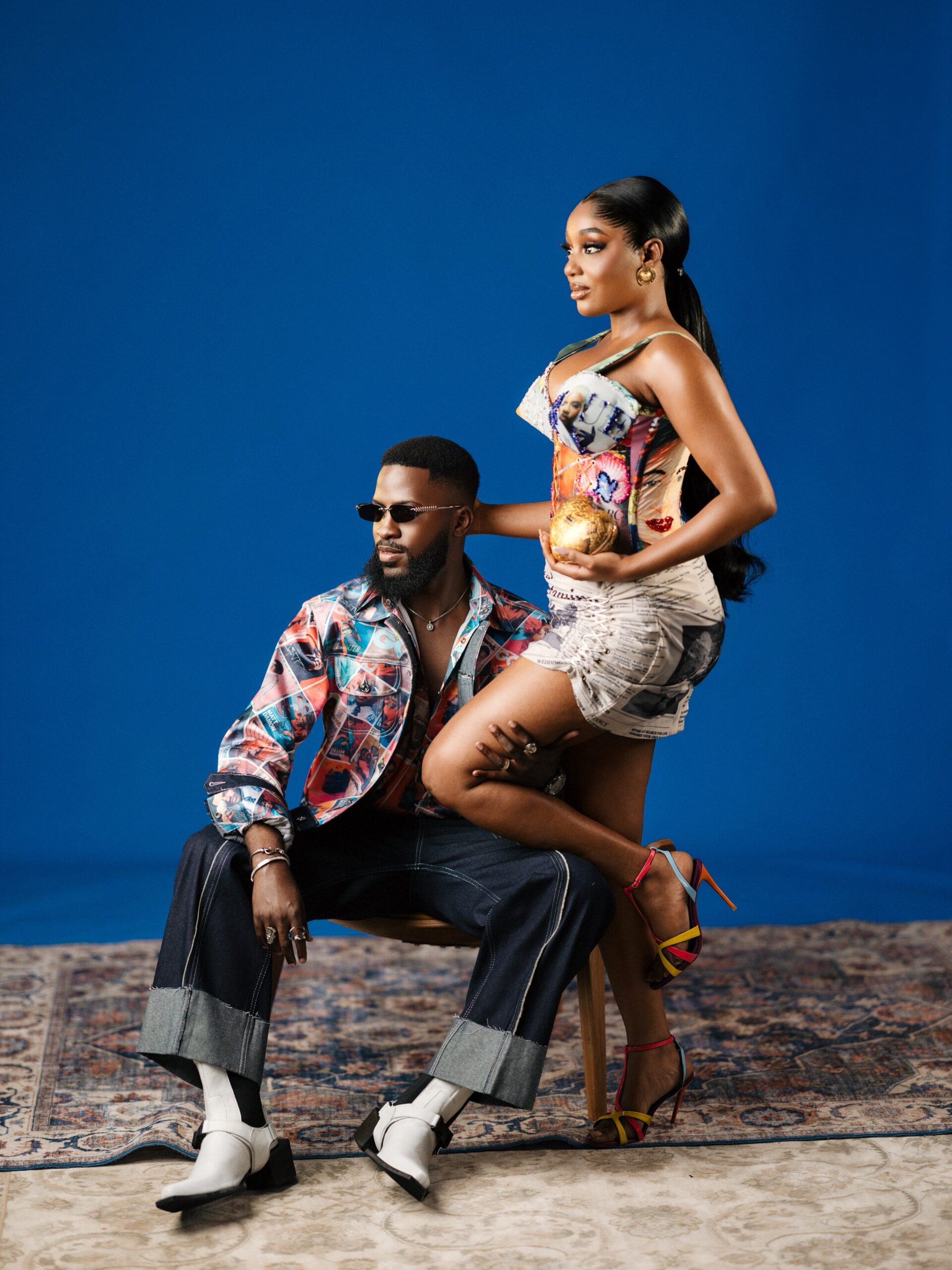 Akin Faminu seated in dark trousers and a graphic shirt with Kiky Festus posing in a Vogue magazine print mini dress.