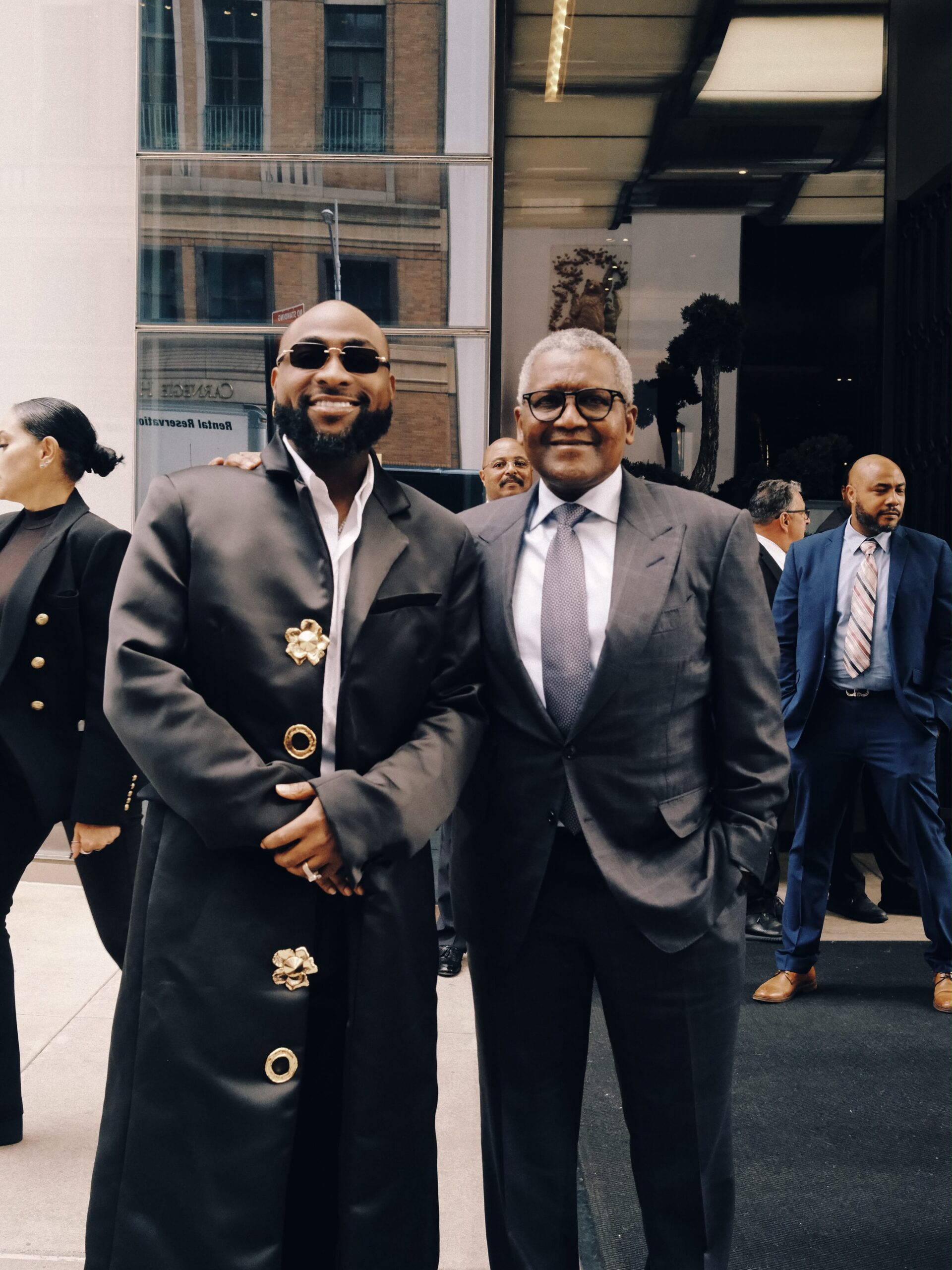 Davido smiling alongside Aliko Dangote in a tailored grey suit with security staff in the background