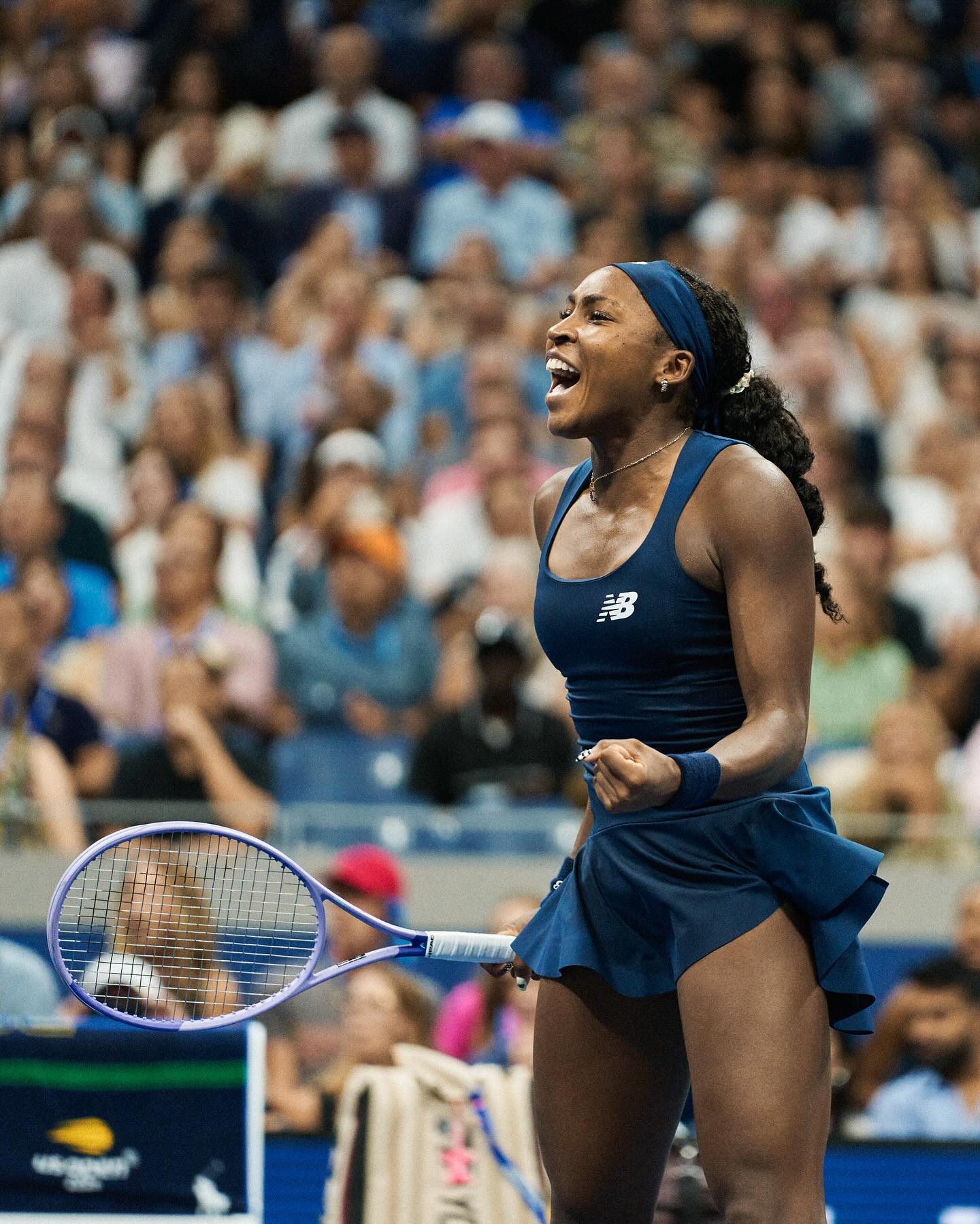 Coco Gauff shouting in celebration with a clenched fist wearing a navy New Balance tennis dress.