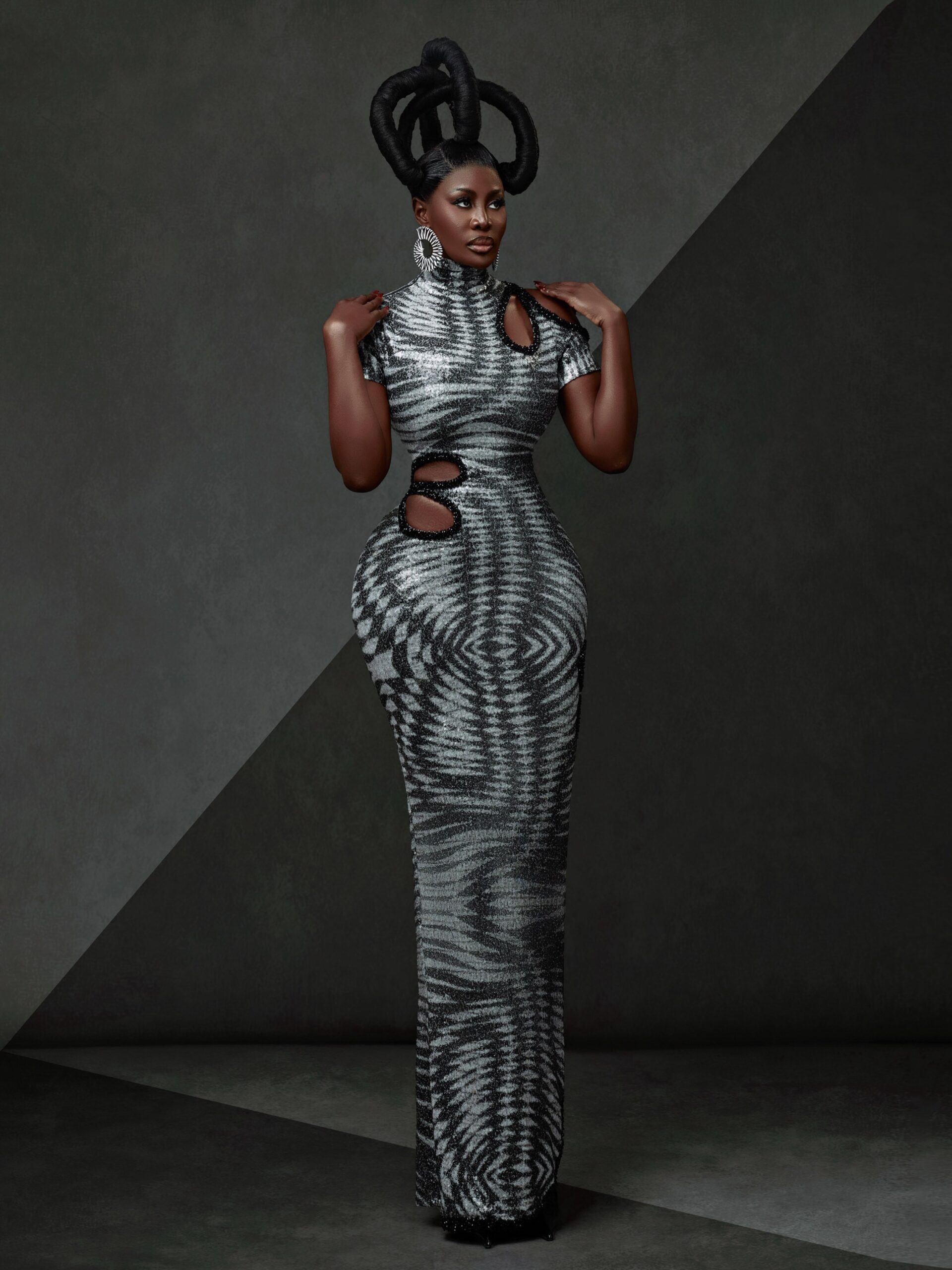 Nana Akua Addo posing in a floor-length grey and black optical-print column gown featuring asymmetrical shoulder and waist cutouts paired with a looped black crown hairstyle.