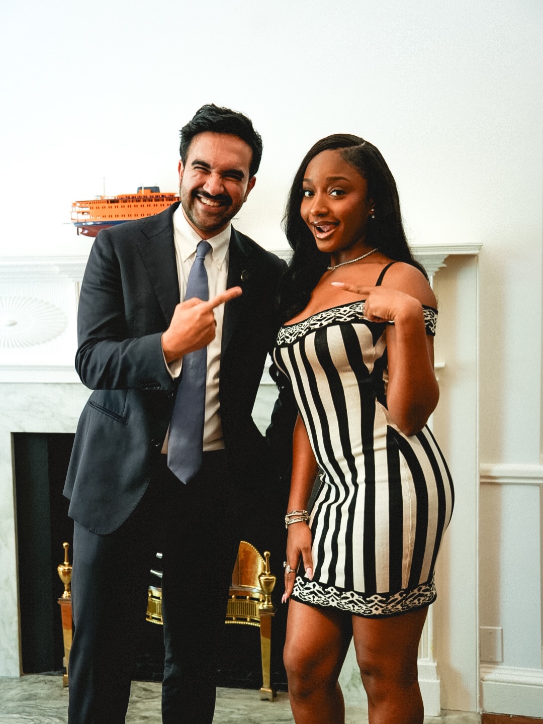 Ayra Starr in a striped knit mini dress and Zohran Mamdani in a dark suit pointing at each other in front of a white fireplace.