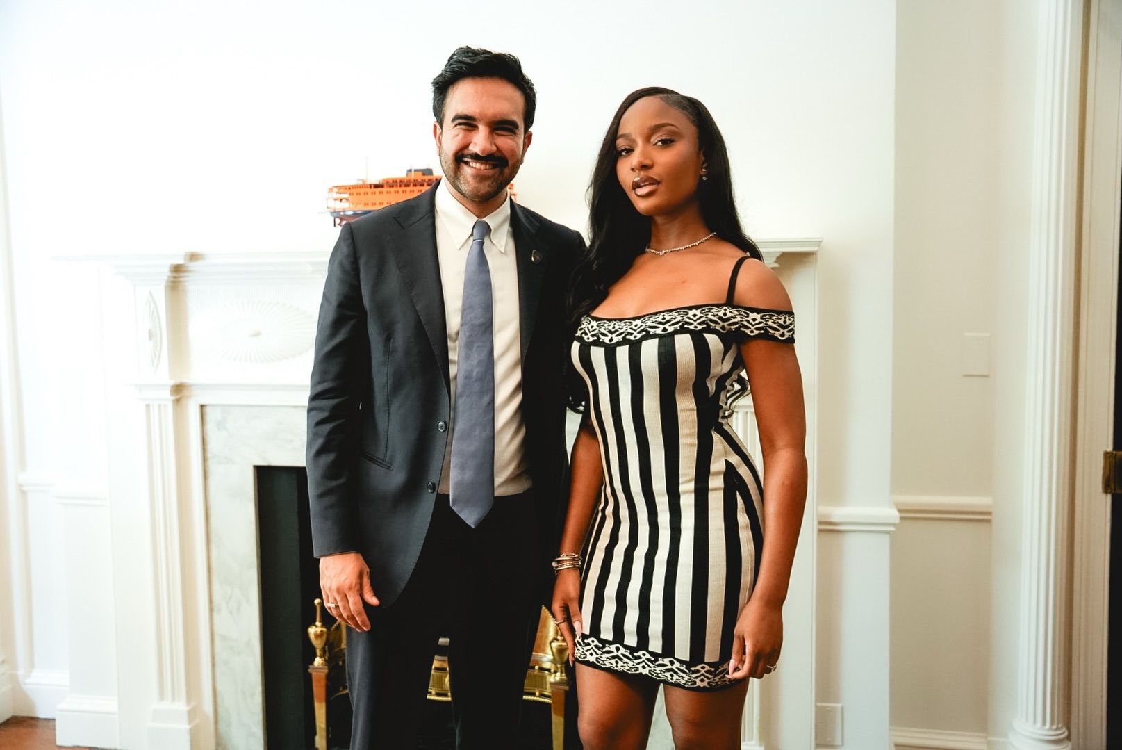 Ayra Starr in a black-and-cream striped dress standing beside Zohran Mamdani in a suit and slate-blue tie in New York.