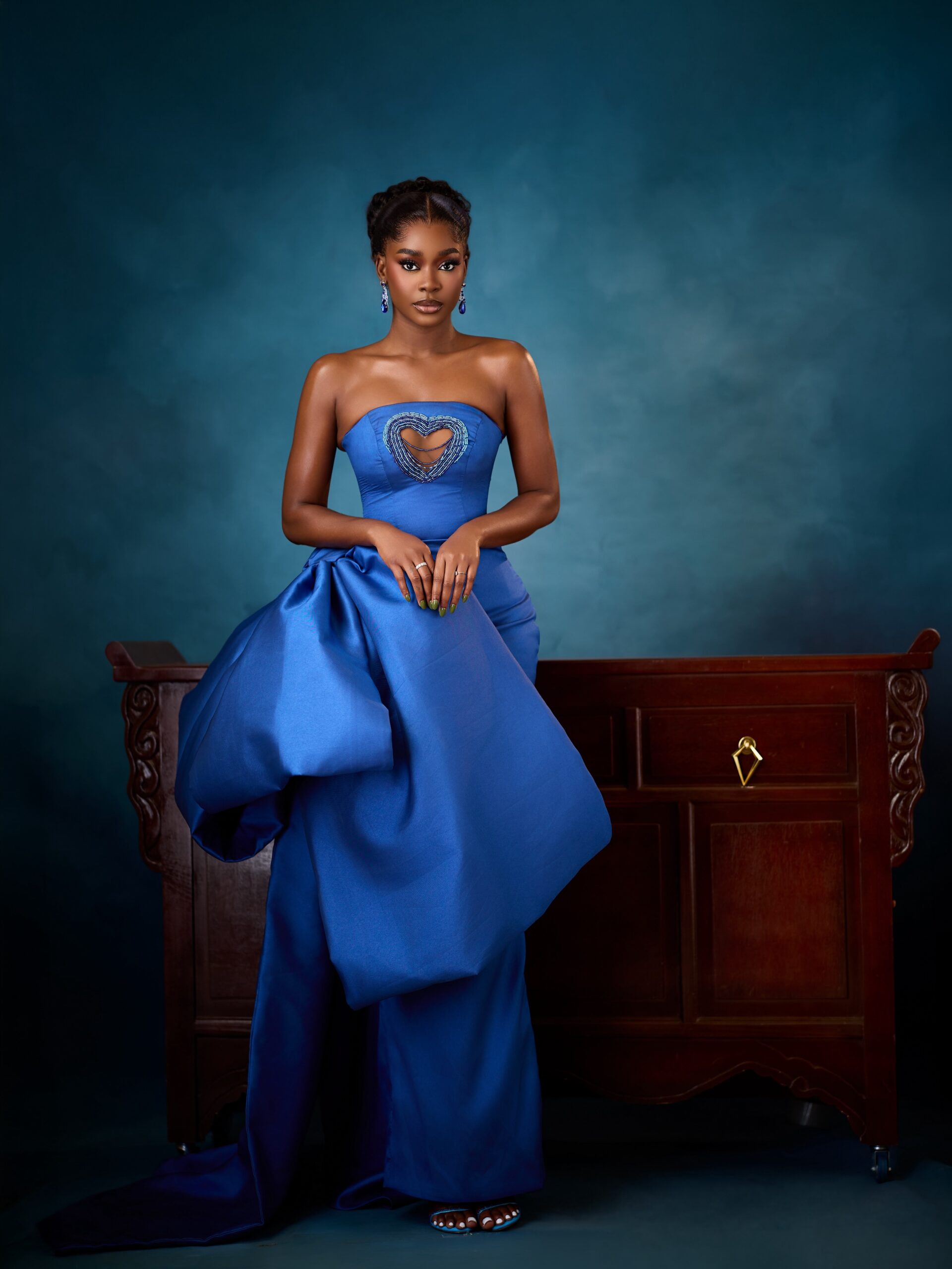 Dede Ashiogwu standing in a royal blue strapless gown with a voluminous hip drape and side train for a wedding guest look.