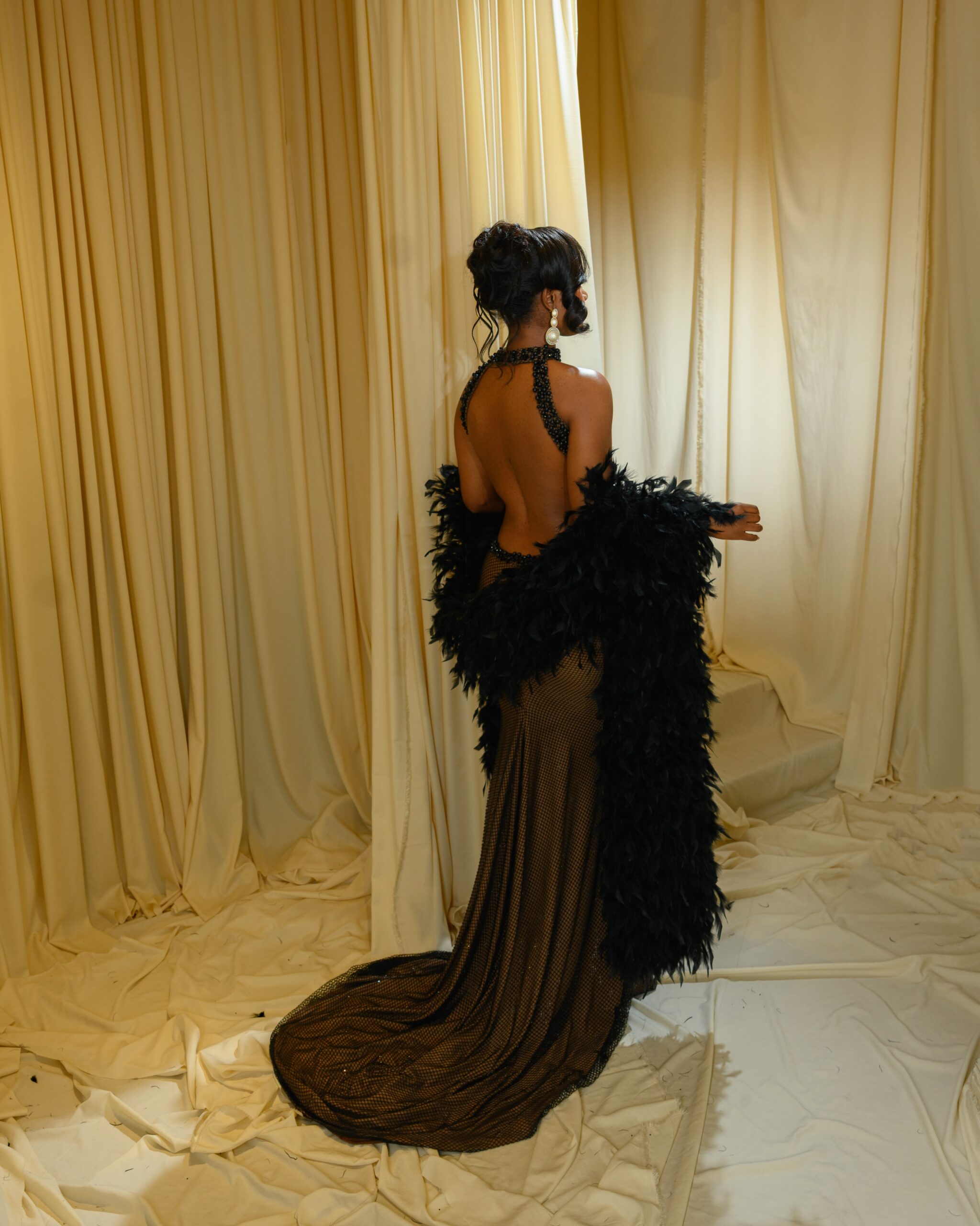 Back view of Bella Okagbue in a backless black fishnet dress with a subtle train and a black feather boa.