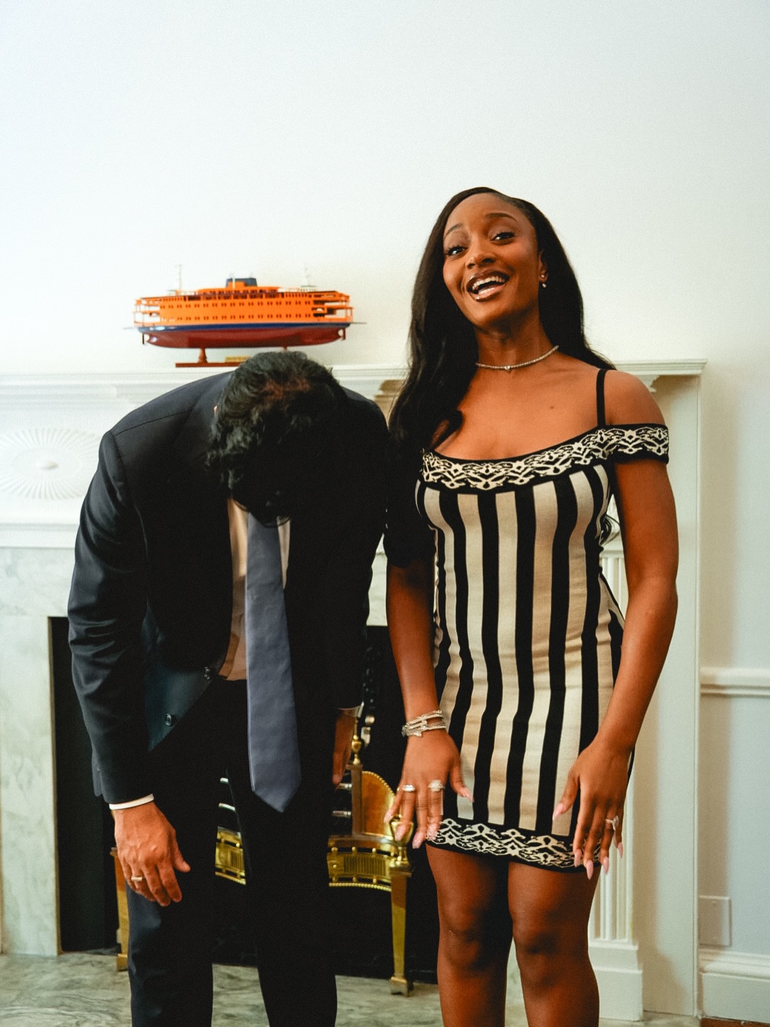 Zohran Mamdani bent over laughing next to Ayra Starr as she smiles in a striped off-the-shoulder knit dress in New York.