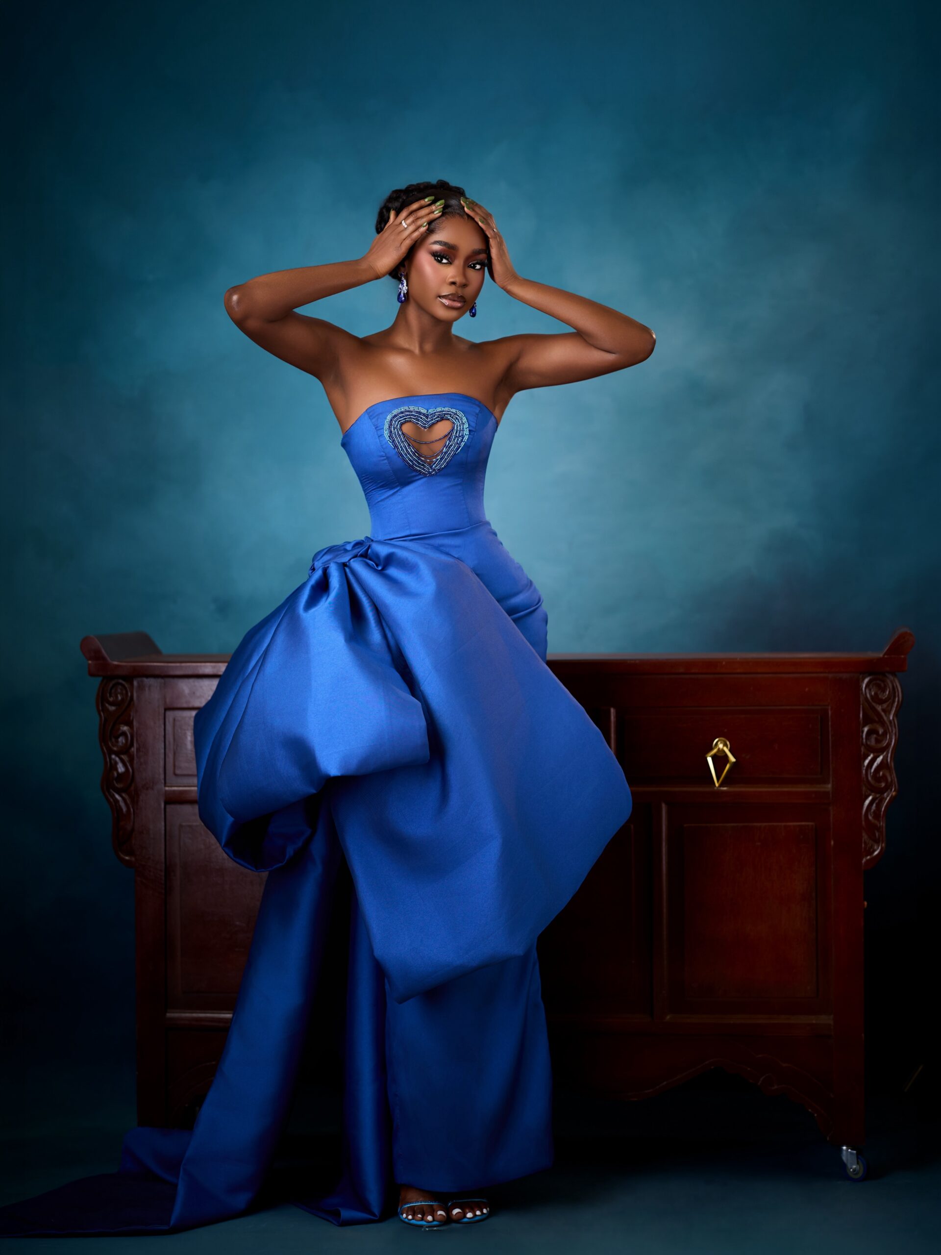 Dede Ashiogwu posing with hands on her head wearing a royal blue strapless gown with a beaded heart cutout.
