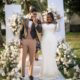 Stan Nze and Blessing Obasi Nzediegwu smiling and showing wedding rings at white floral altar during vow renewal.
