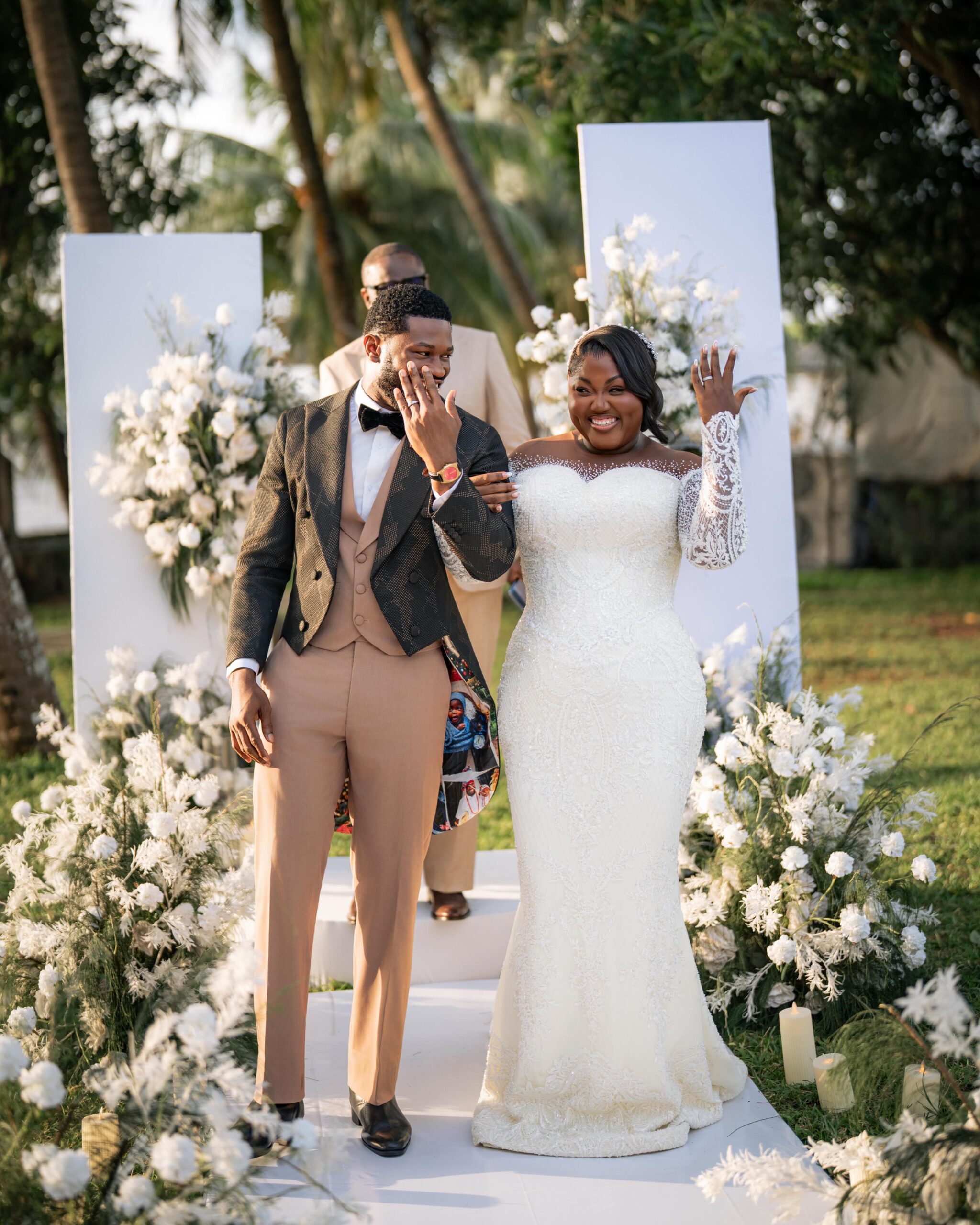 Stan Nze and Blessing Obasi Nzediegwu smiling and showing wedding rings at white floral altar during vow renewal.