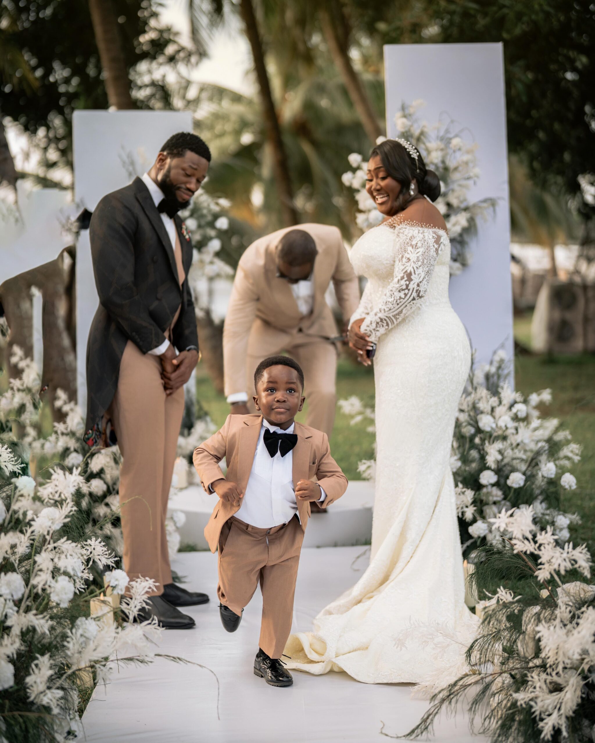 Jayden Nze running happily down white aisle in tan suit while parents Stan Nze and Blessing Obasi Nzediegwu smile at altar.