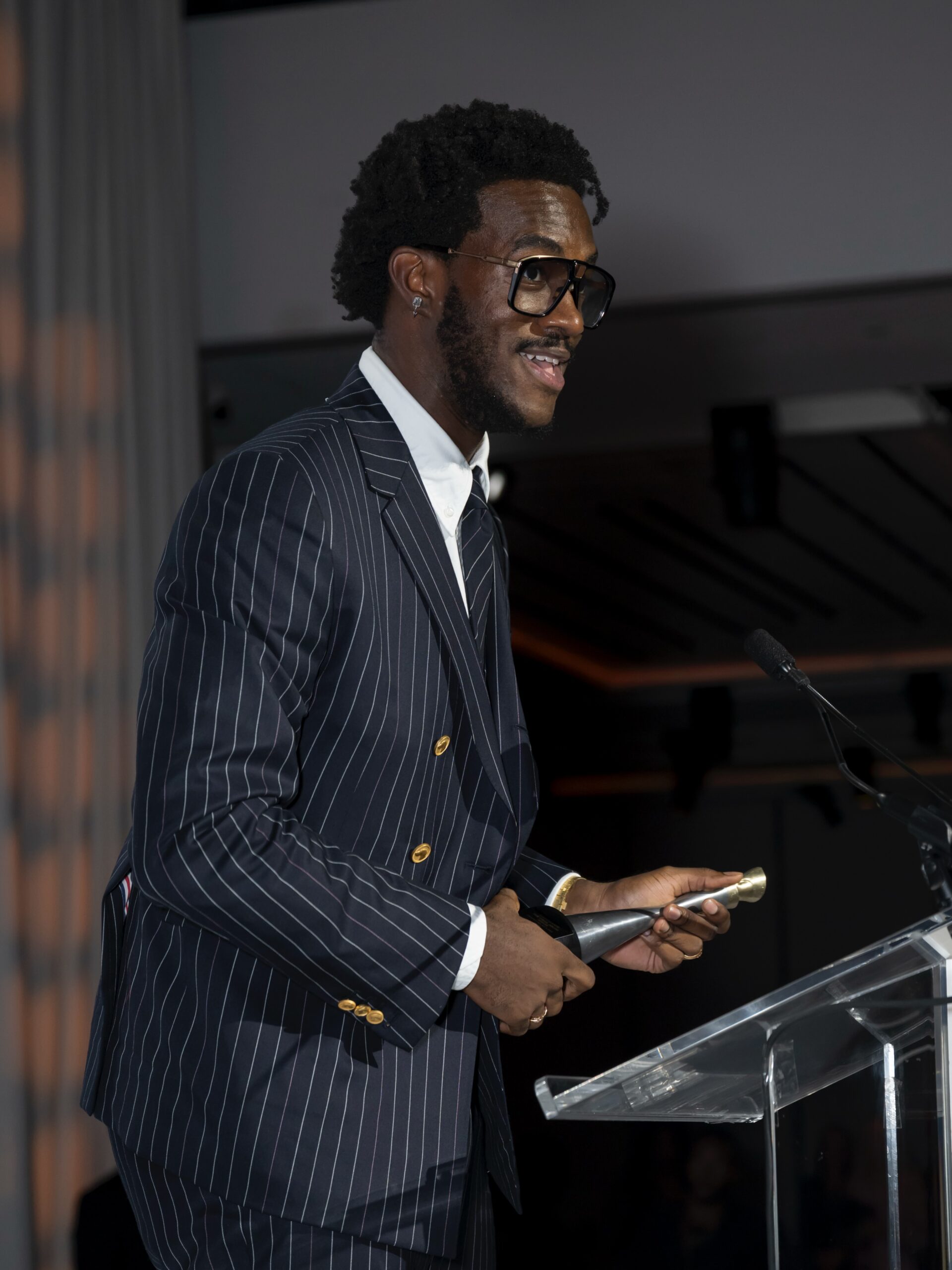 Wisdom Kaye holding silver trophy at podium during acceptance speech for Virgil Abloh Award at Harlem's Fashion Row.