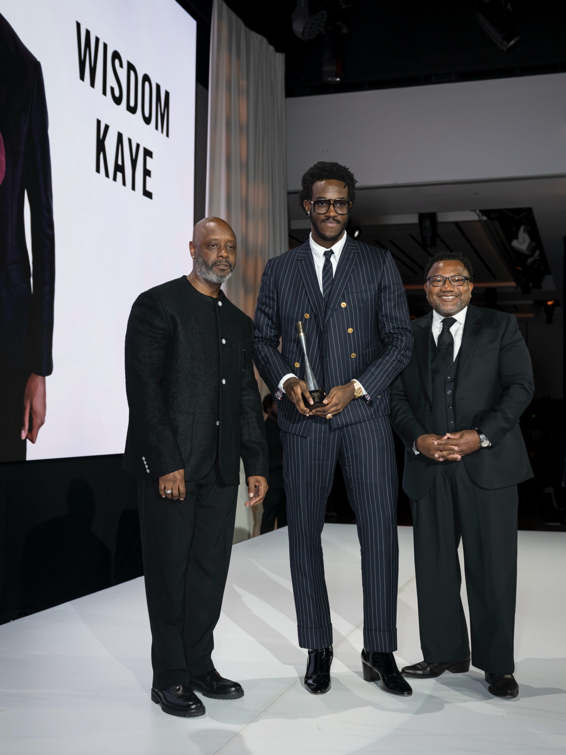 Wisdom Kaye standing on stage with trophy alongside LVMH executives at Harlem's Fashion Row Style Awards.