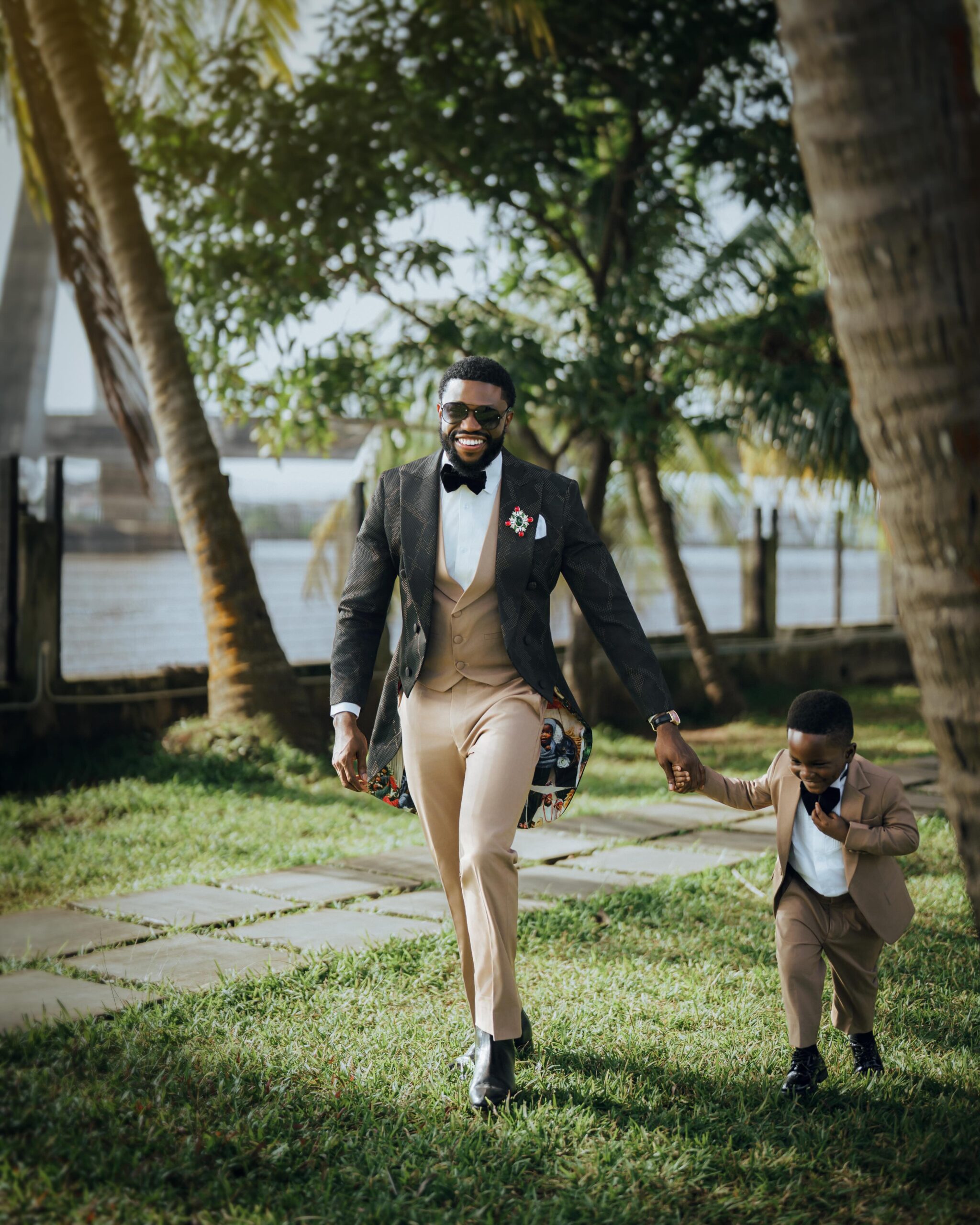 Actor Stan Nze in P.Aries suit walking holding hands with his young son Jayden in matching tan outfit at vow renewal ceremony.