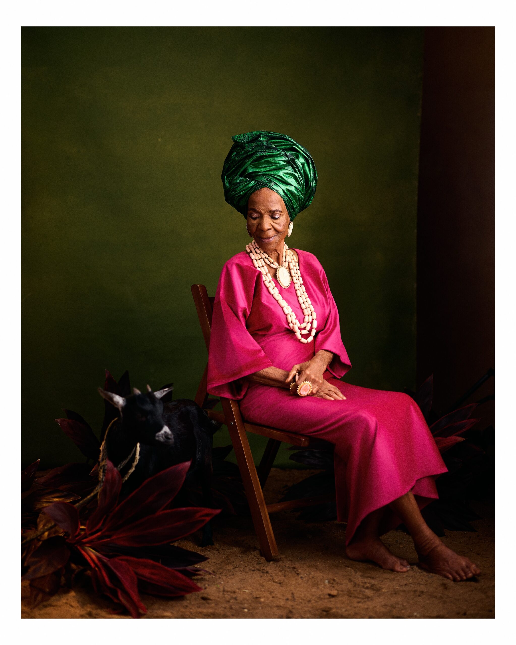 Veteran Yoruba actress Grace Owoola, famous as Iya Oshogbo, wearing a hot-pink buba and iro, and emerald green gele next to a black goat for her 96th birthday photoshoot.