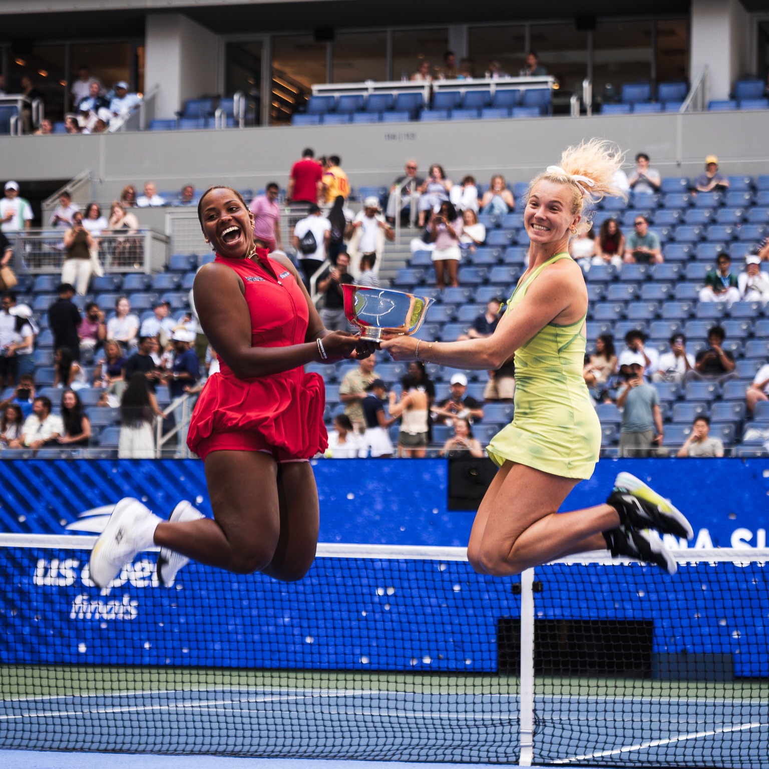 Taylor Townsend and Katerina Siniakova jumping mid-air holding the US Open trophy.