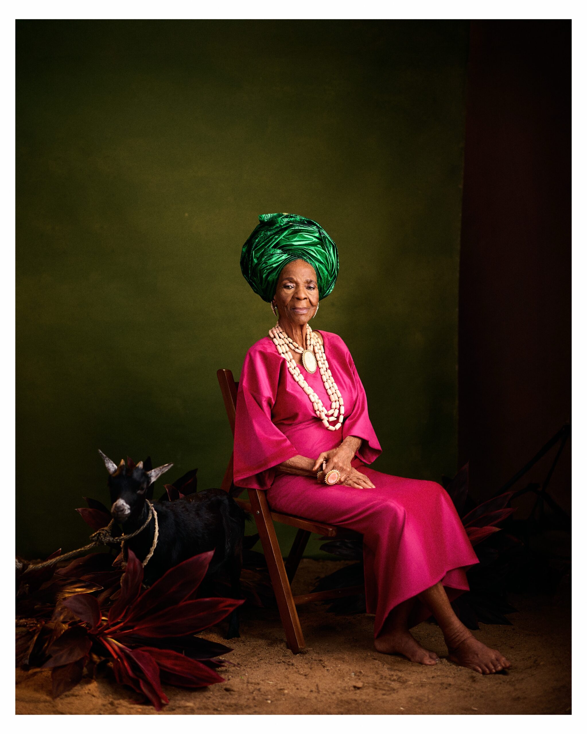 Grace Owoola looking directly at the camera in a pink gown, high green gele, and coral beads seated beside a black goat.