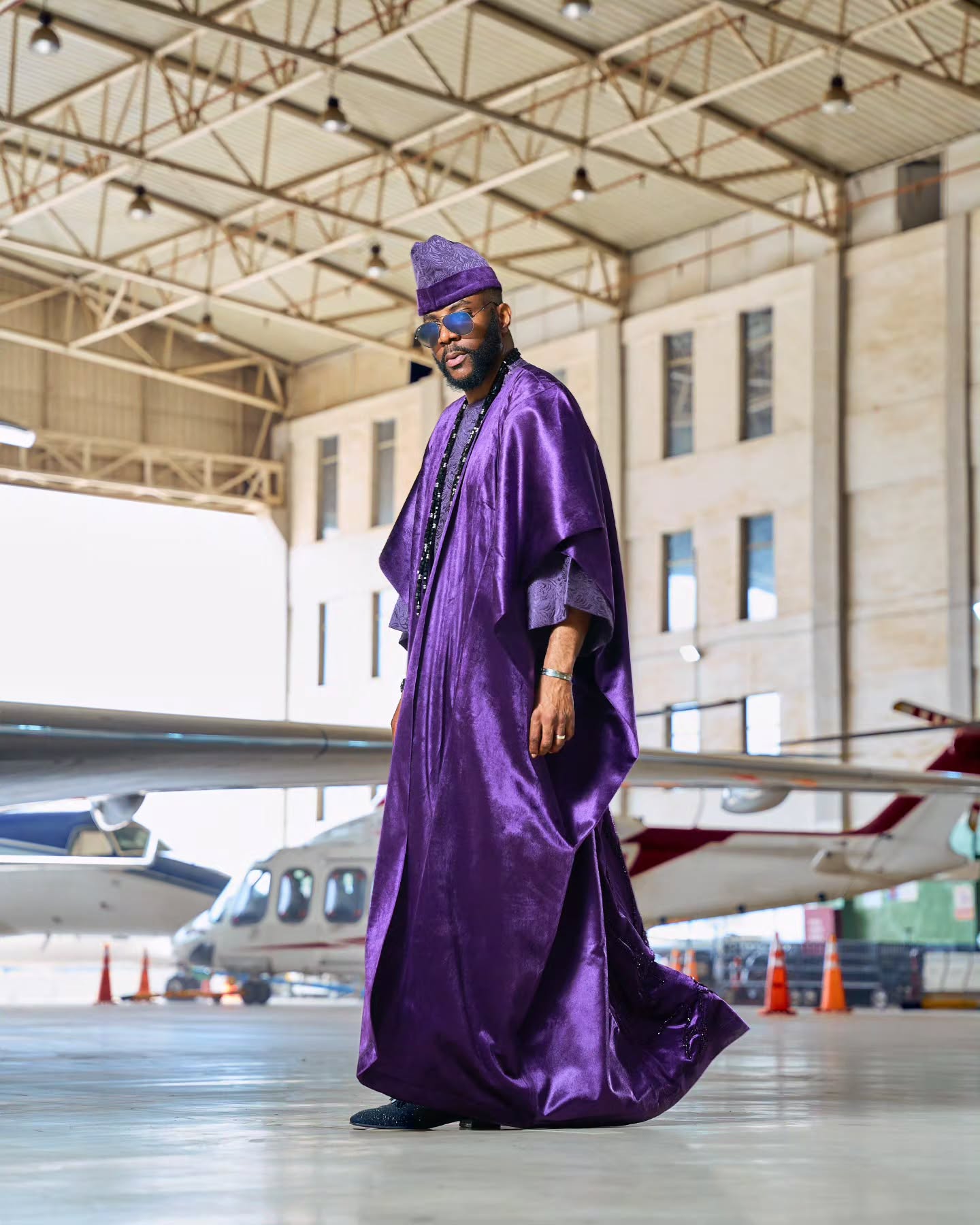 Ebuka Obi-Uchendu posing in a purple velvet agbada by D29 Africa inside an airfield hangar with private jets.