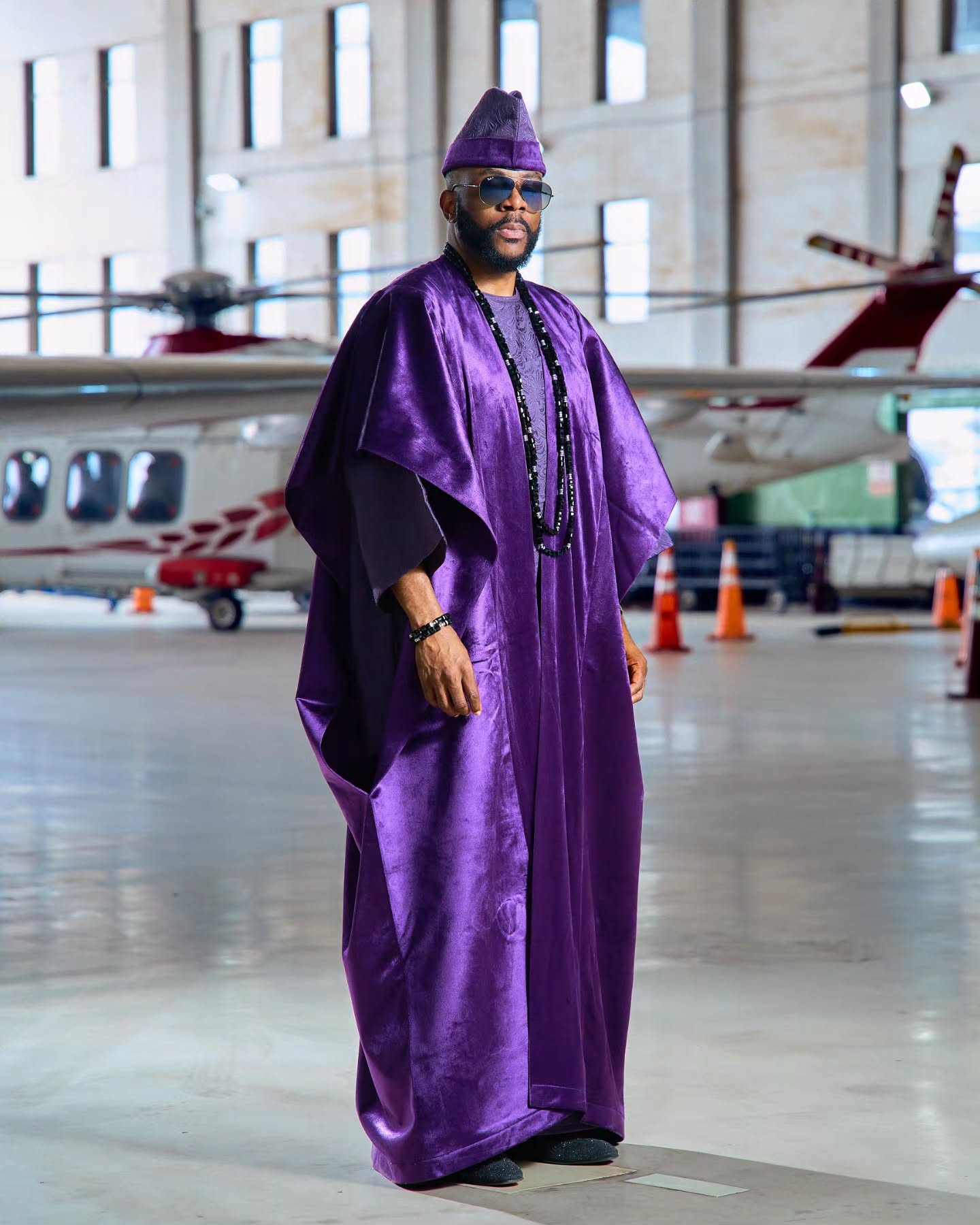 Ebuka Obi-Uchendu standing inside a hangar wearing a purple velvet agbada and matching fila by D29 Africa.