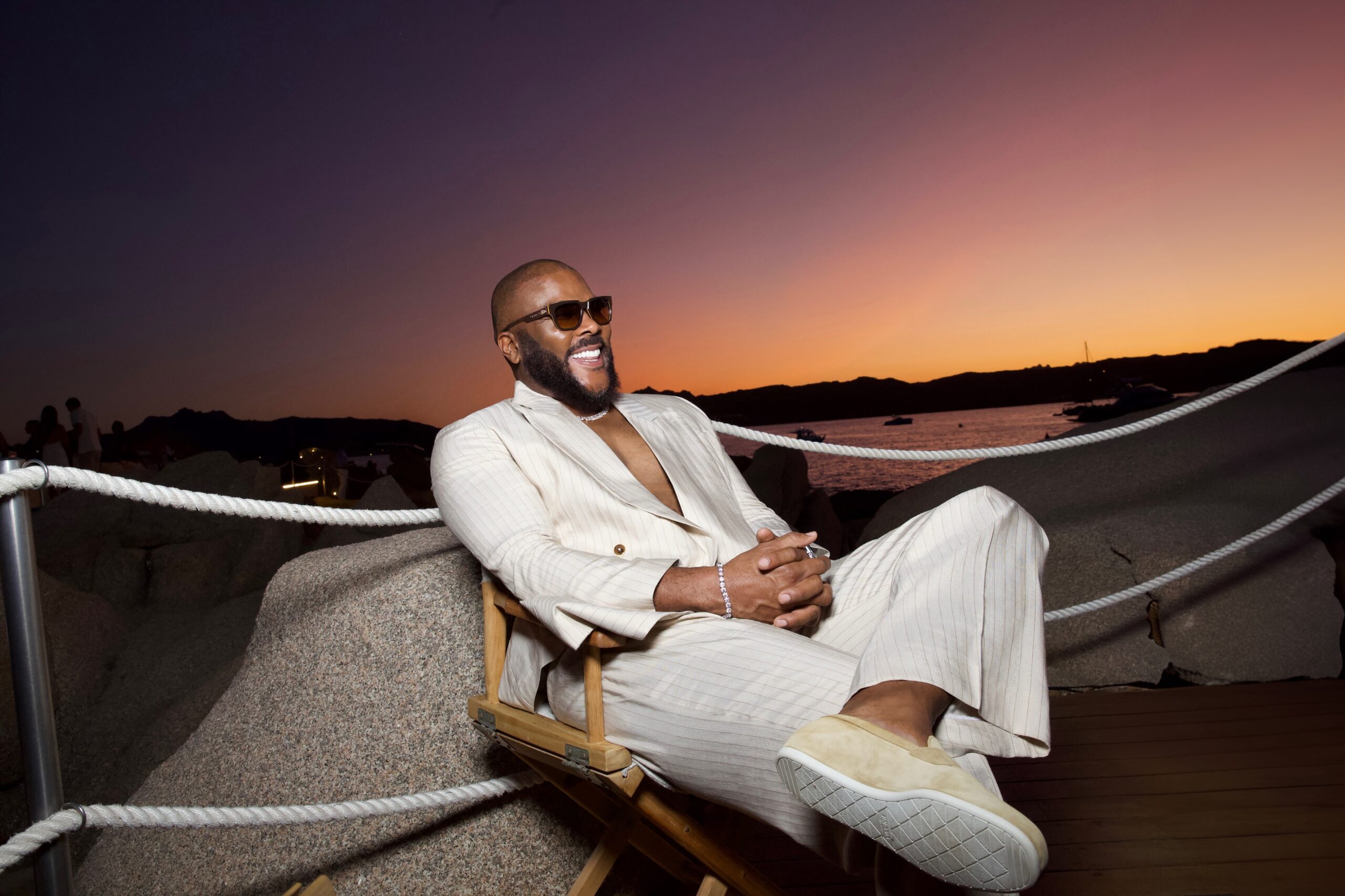 Tyler Perry celebrating his 57th birthday in a pinstripe cream suit and sunglasses against a sunset background.