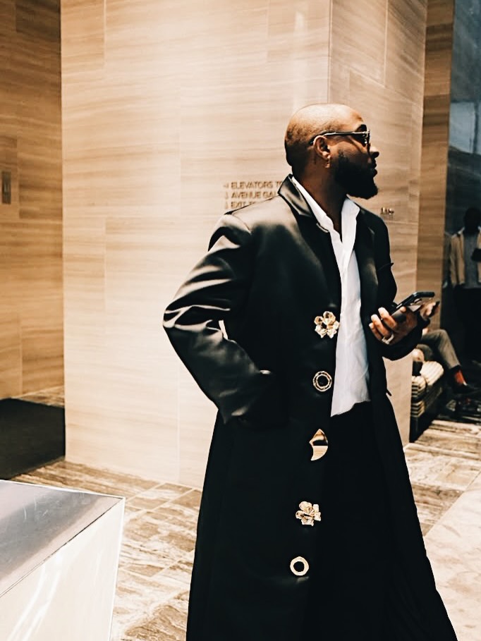 Davido holding a smartphone in a floor-length black duster coat with gold metallic hardware buttons in a New York hotel lobby
