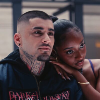 Ayra Starr resting her arm on Zayn Malik's shoulder as he wears a dark embroidered bomber jacket with neck tattoos visible.