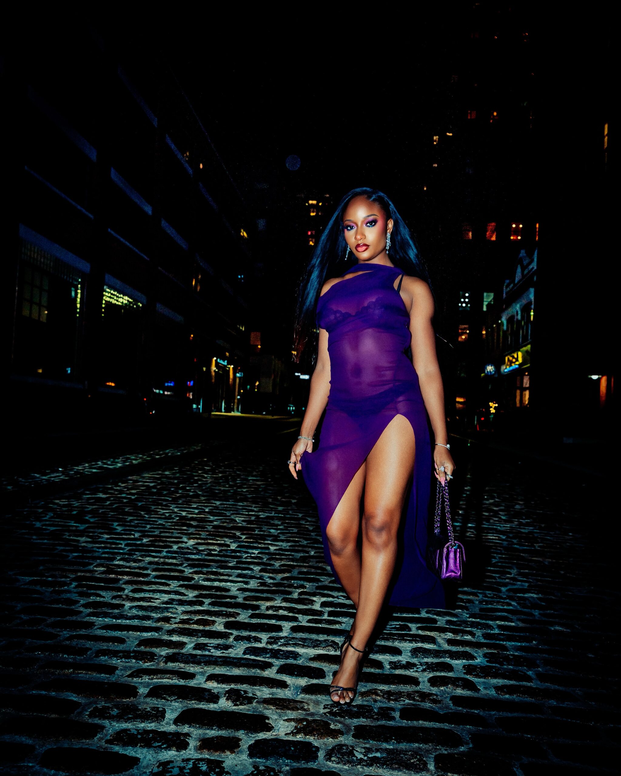 Ayra Starr walking along a cobblestone street at night in a royal-purple semi-sheer high-slit dress by LaQuan Smith with black heels.