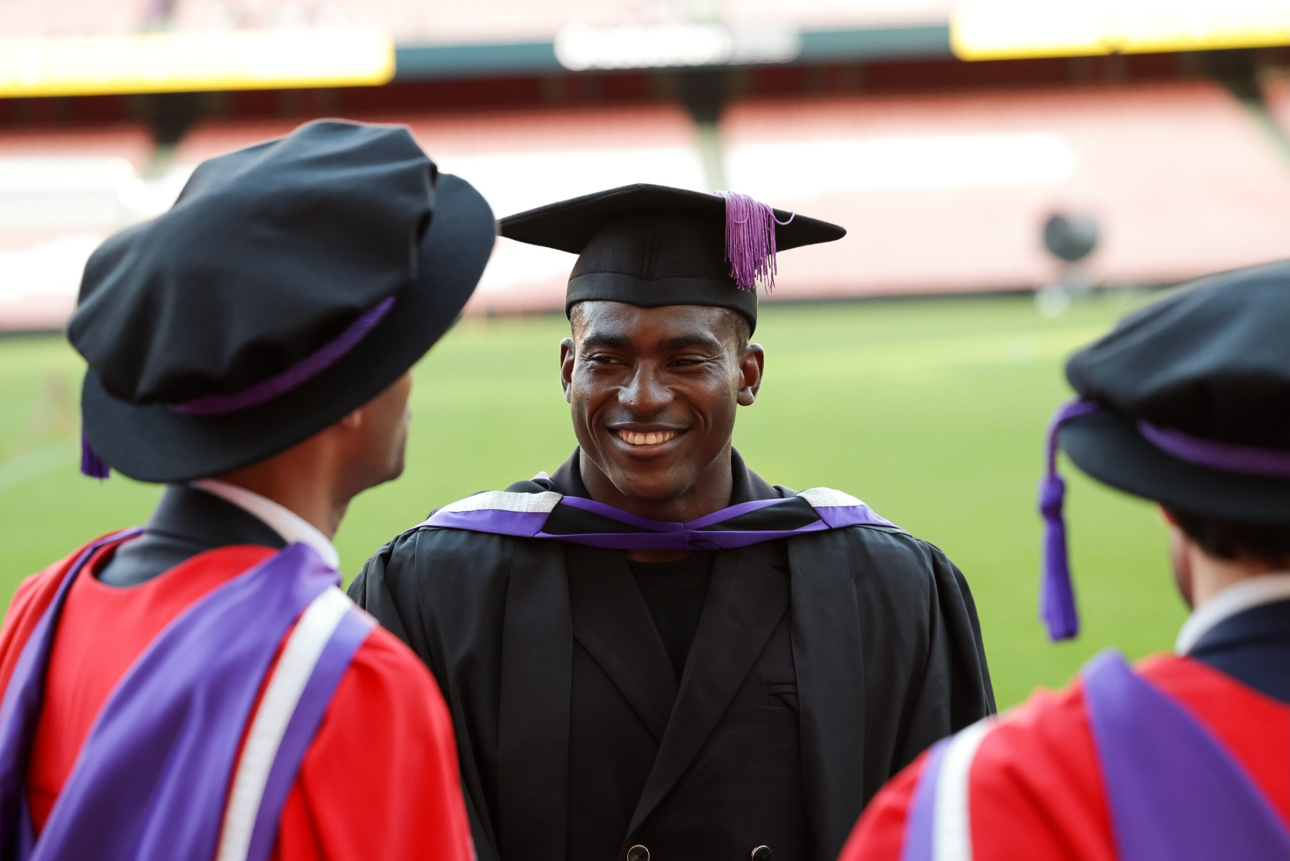 Taiwo Awoniyi wearing a cap and gown smiling during a conversation with fellow graduates at a football stadium
