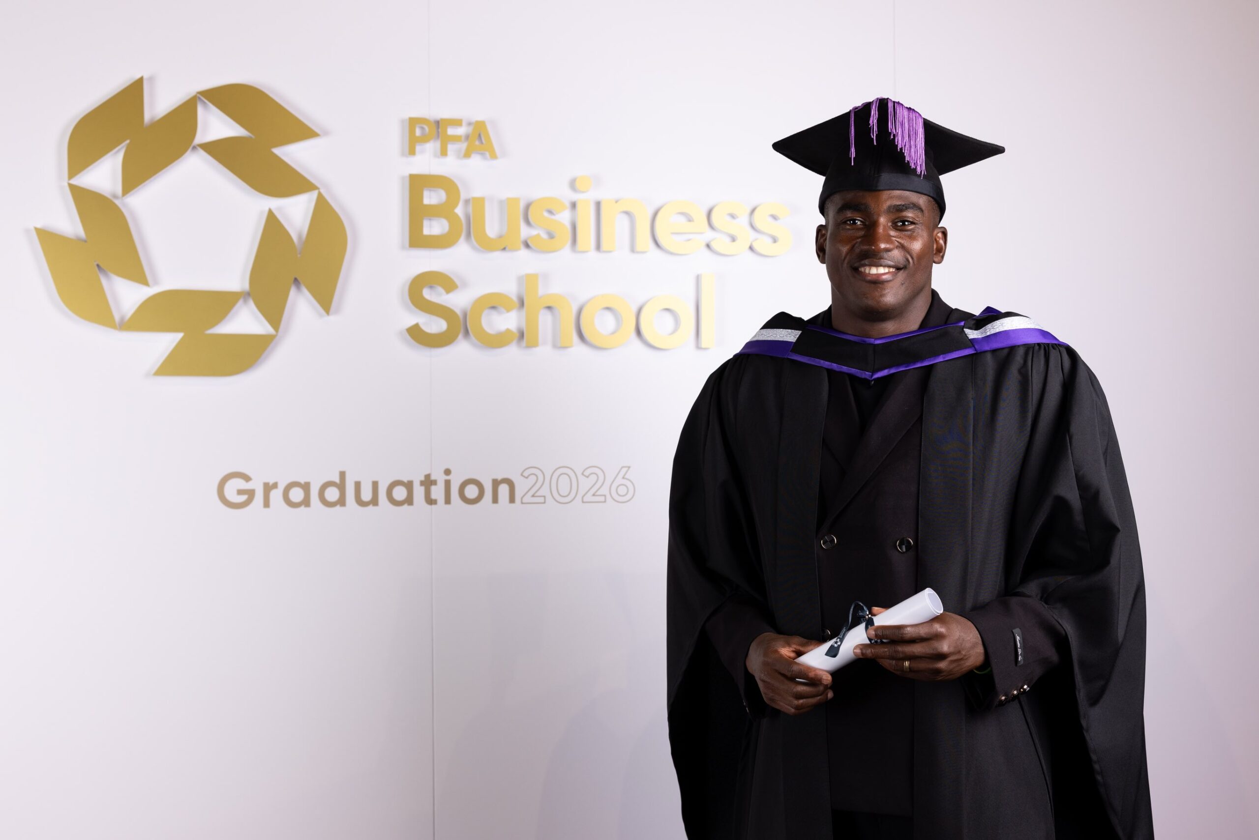 Taiwo Awoniyi smiling in academic cap and gown holding his diploma scroll in front of the PFA Business School graduation logo