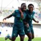 Two Nigeria Falconets players smiling and running side-by-side with arms around each other to celebrate a goal.