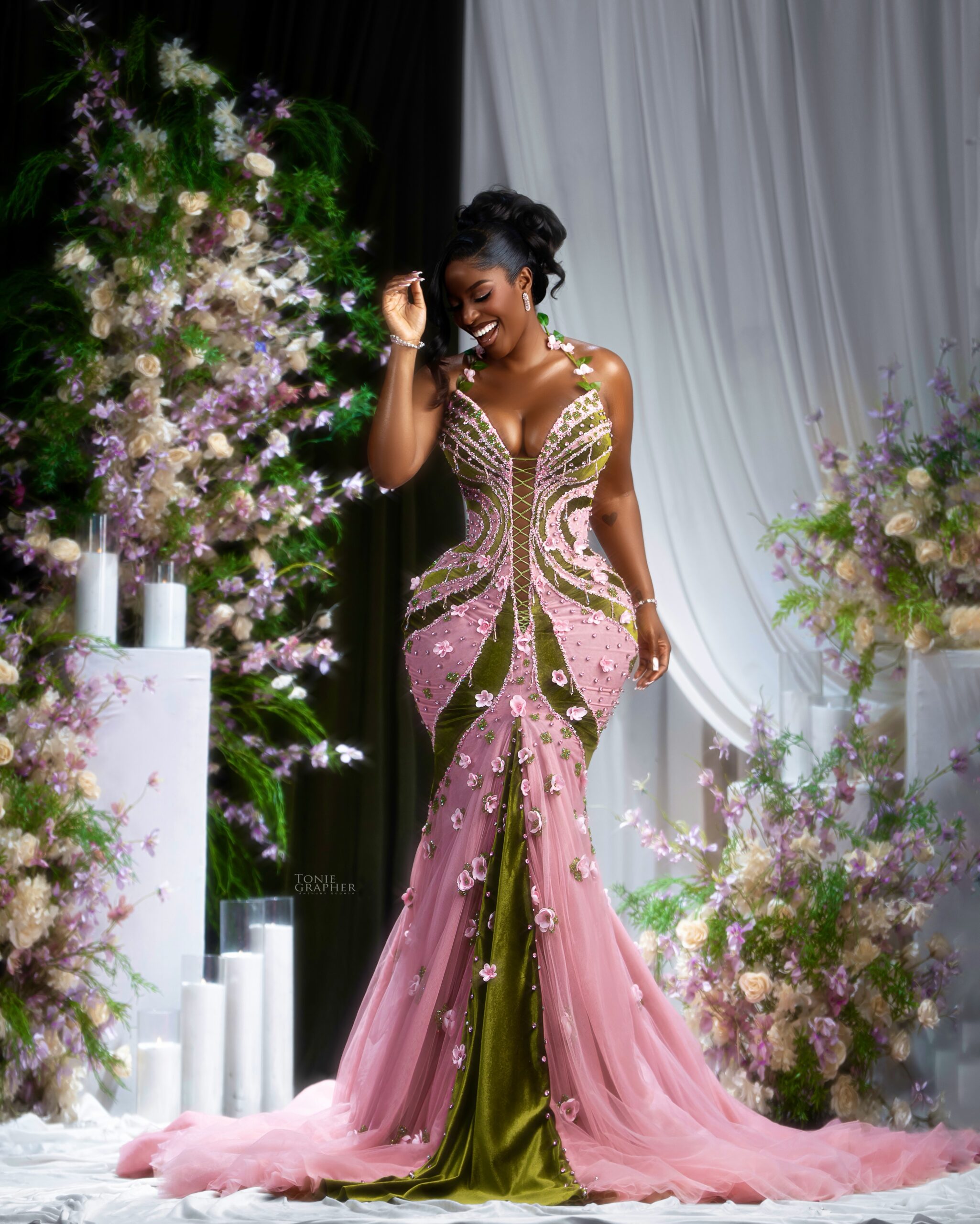 Hilda Baci smiling in an olive green and blush pink floral corseted mermaid gown amidst floral arrangements.