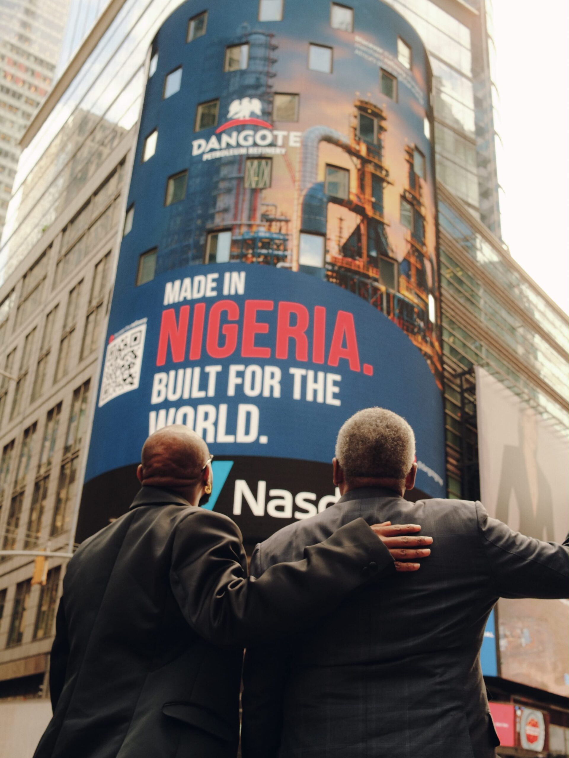 Back-facing view of Davido and Aliko Dangote with arms around each other looking up at the Dangote Refinery Nasdaq billboard in Times Square reading Made in Nigeria Built for the World