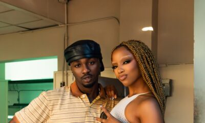 Omah Lay and Chlöe Bailey standing together in a corridor; Omah Lay in a striped polo and baker boy cap, Chlöe in a knit bodysuit.