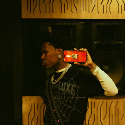 Shoday holding a phone displaying the title of his song CHE in a music studio
