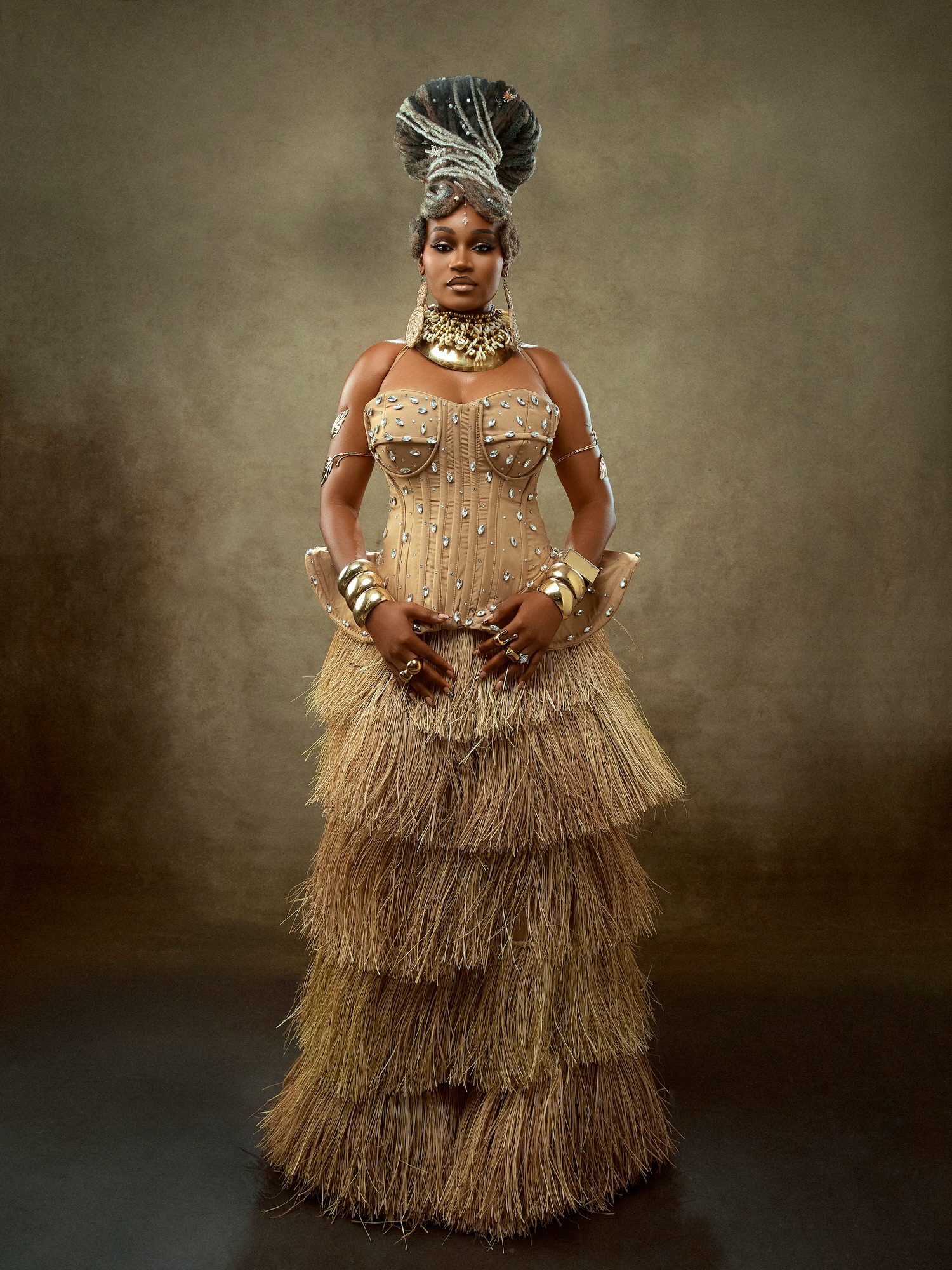 J’Dess standing in a floor-length tiered raffia skirt, crystal-studded corset, and sculpted locs headpiece for her ALA EP cover styling.