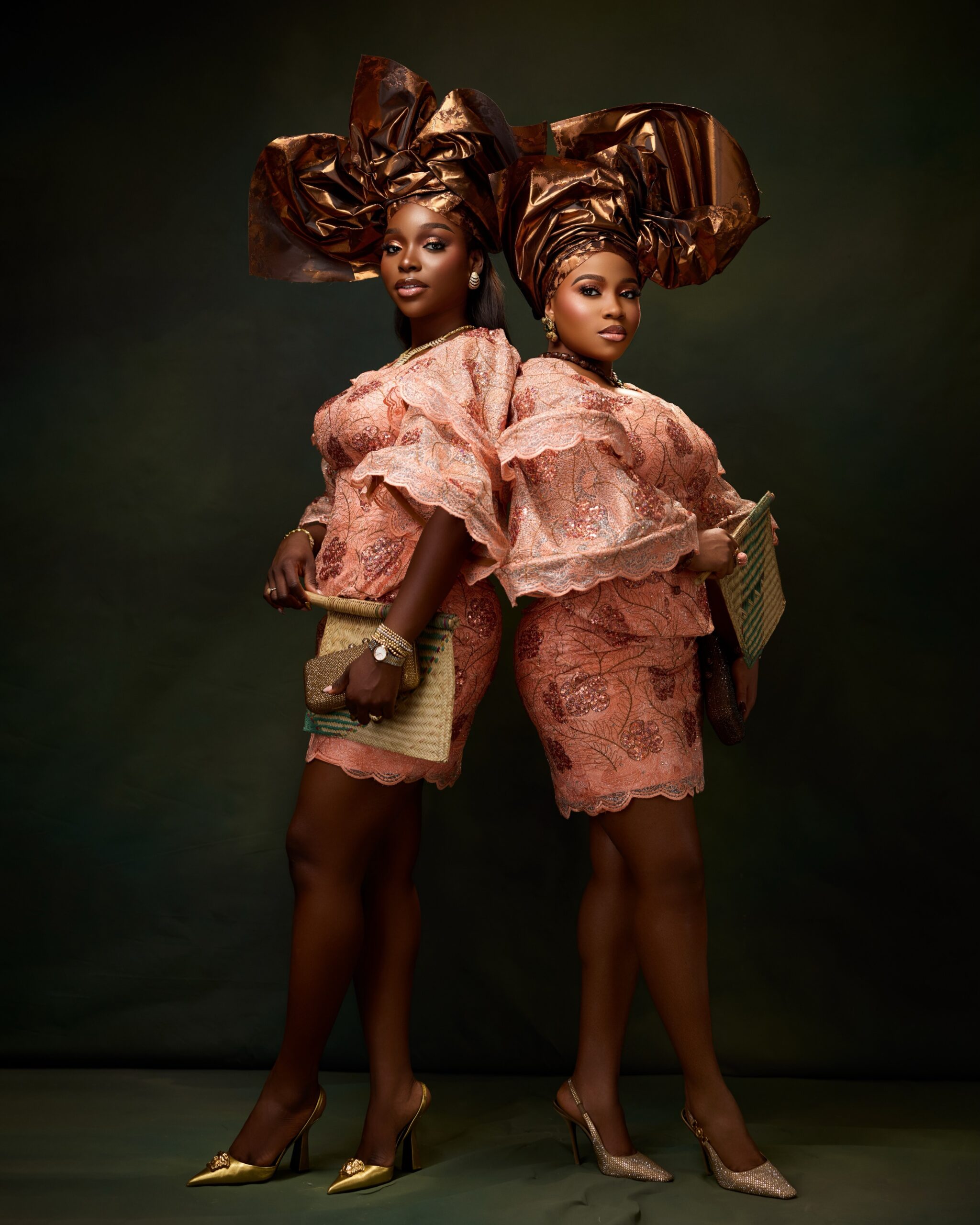 Veekee James and Tomike Adeoye standing back to back wearing matching peach sequin lace skirt and blouse sets with bronze-gold structural headties