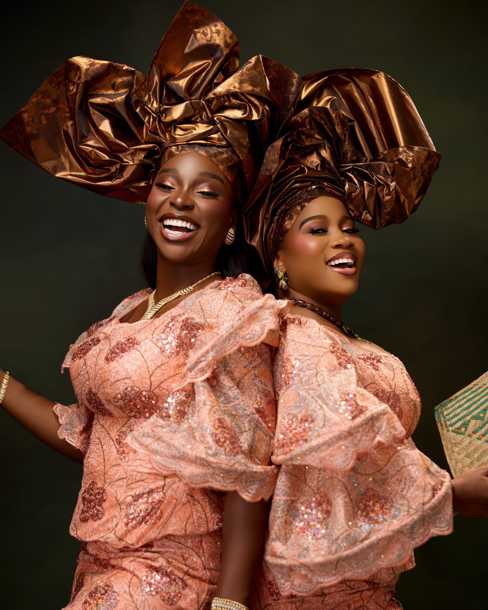 Veekee James and Tomike Adeoye smiling in peach sequin lace blouses, bronze-gold metallic headties, and warm bronzed makeup