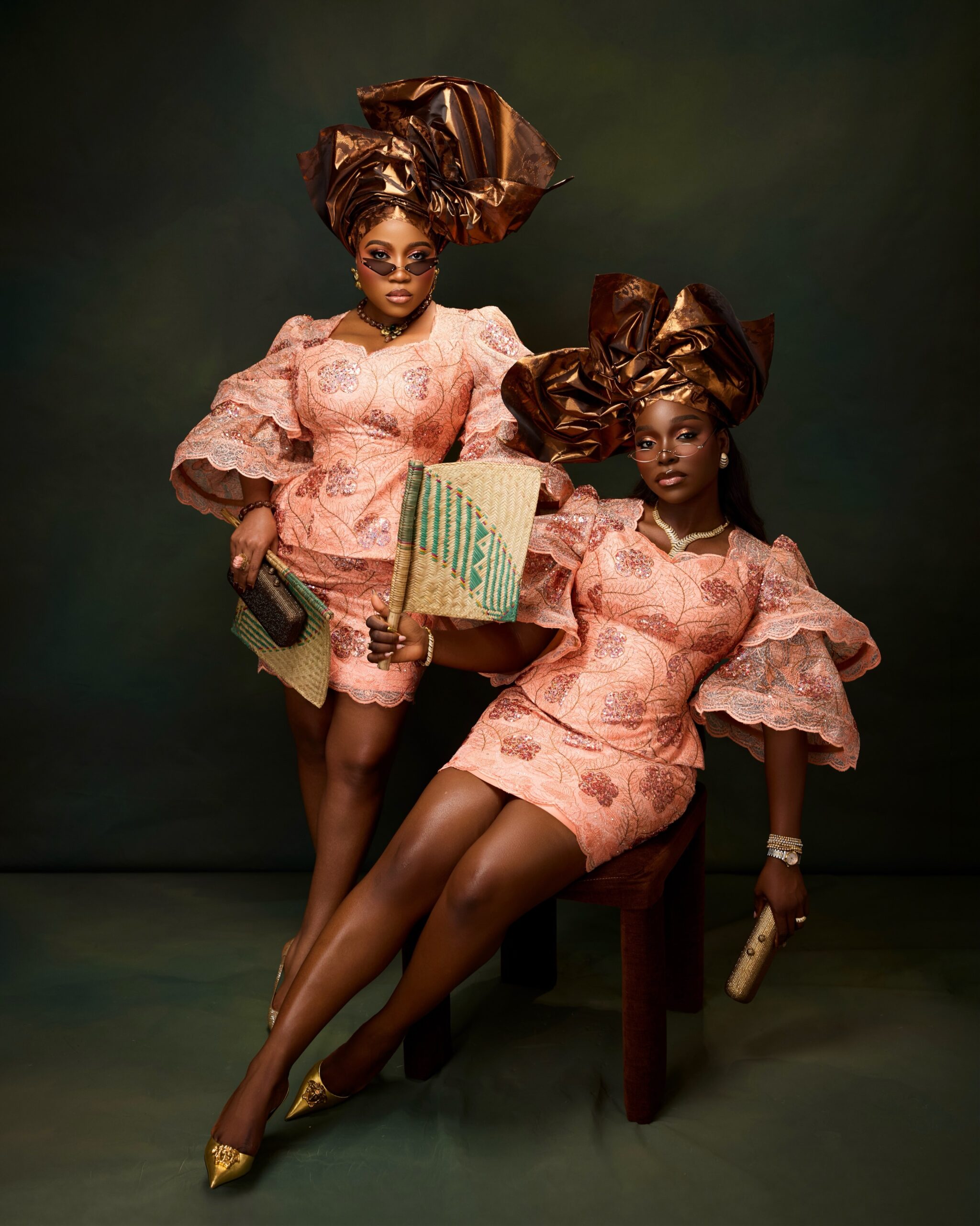 Veekee James seated and Tomike Adeoye standing wearing dark sunglasses, matching peach lace sets, and bronze-gold headties