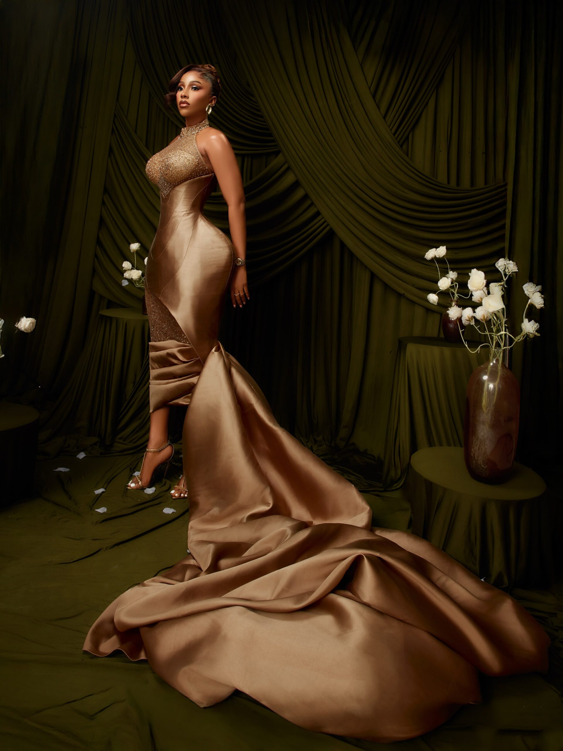 Mercy Eke posing in a high-neck champagne-bronze gown with sheer crystal bodice detailing and a dramatic draped satin skirt.