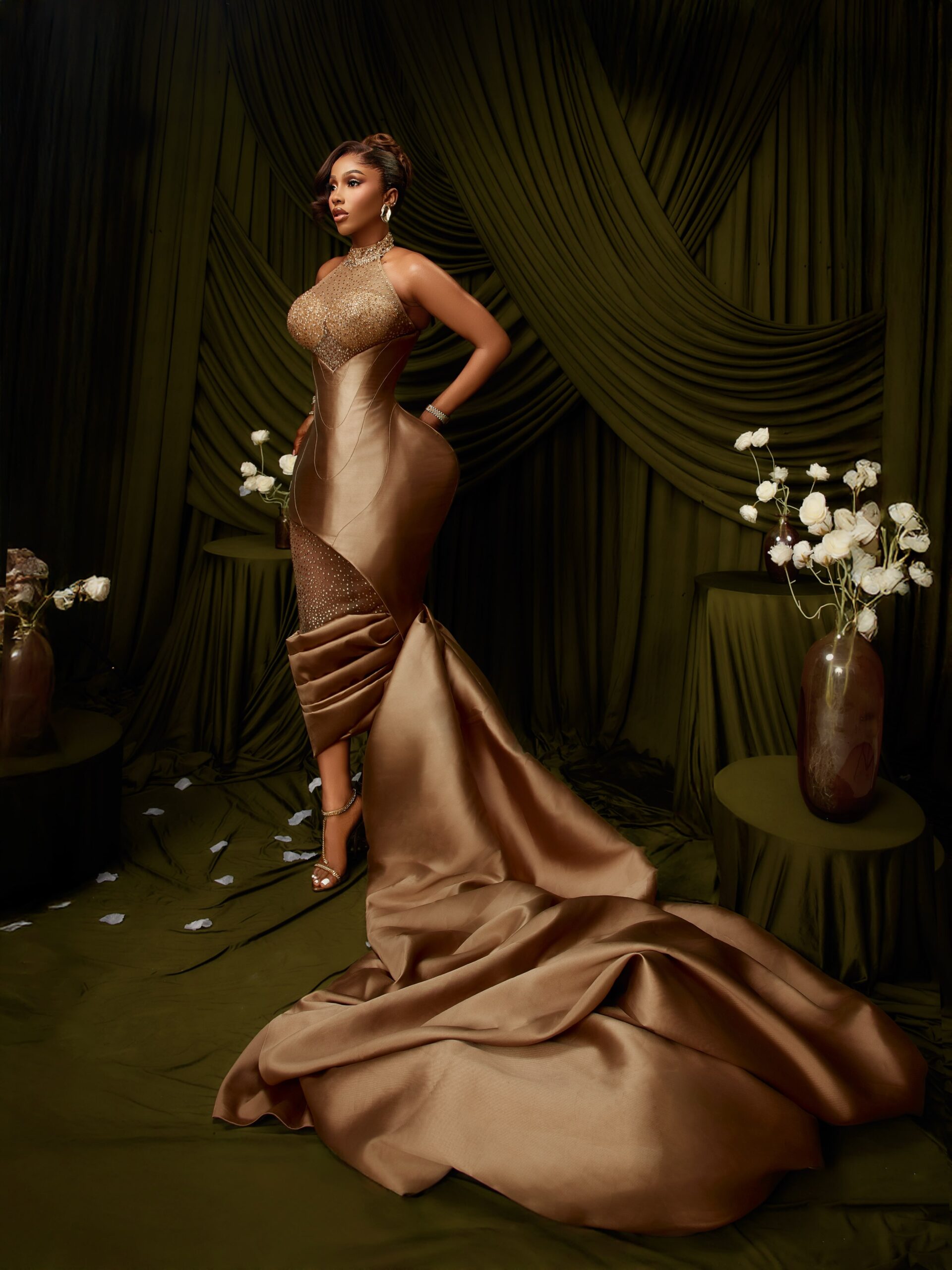 Mercy Eke in a champagne-bronze evening dress with an exaggerated hip corset, asymmetric knee draping, and a long silk train.