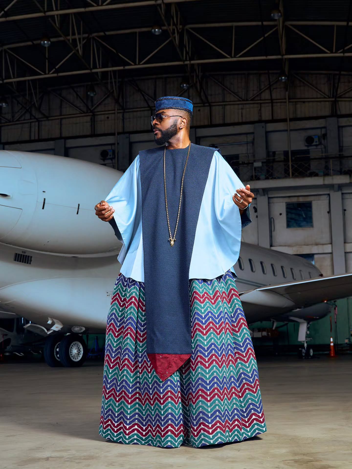 Ebuka Obi-Uchendu wearing a custom sky-blue Ugo Monye tunic with flared butterfly sleeves, a textured navy chest panel, and wide-legged chevron trousers in an airfield hangar.