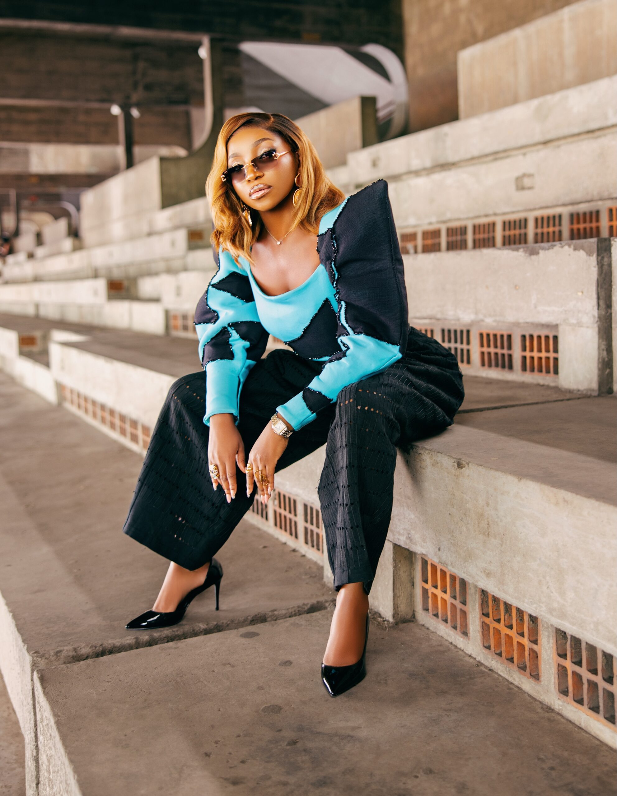 Omowunmi Dada seated on concrete bleachers in a custom turquoise and black outfit at the Black Market Lagos premiere