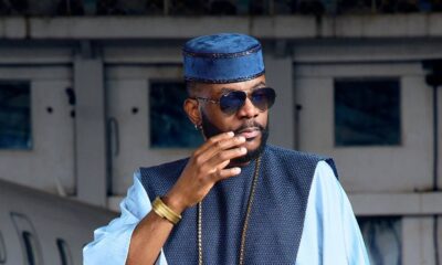 Ebuka Obi-Uchendu adjusting his aviator sunglasses while wearing a sky-blue flared-sleeve tunic and royal blue velvet cap.