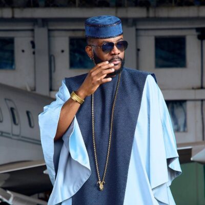 Ebuka Obi-Uchendu adjusting his aviator sunglasses while wearing a sky-blue flared-sleeve tunic and royal blue velvet cap.