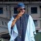 Ebuka Obi-Uchendu adjusting his aviator sunglasses while wearing a sky-blue flared-sleeve tunic and royal blue velvet cap.