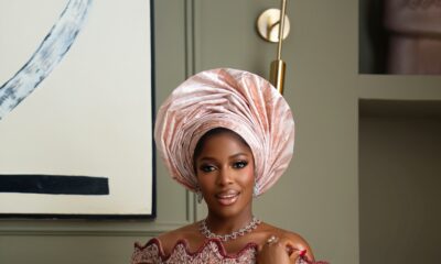 Hilda Baci wearing a peach corseted lace dress by Dorhann with burgundy floral appliqués, a scalloped off-the-shoulder neckline, a tall pleated gele, and holding a wine red satin clutch.