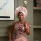 Hilda Baci wearing a peach corseted lace dress by Dorhann with burgundy floral appliqués, a scalloped off-the-shoulder neckline, a tall pleated gele, and holding a wine red satin clutch.