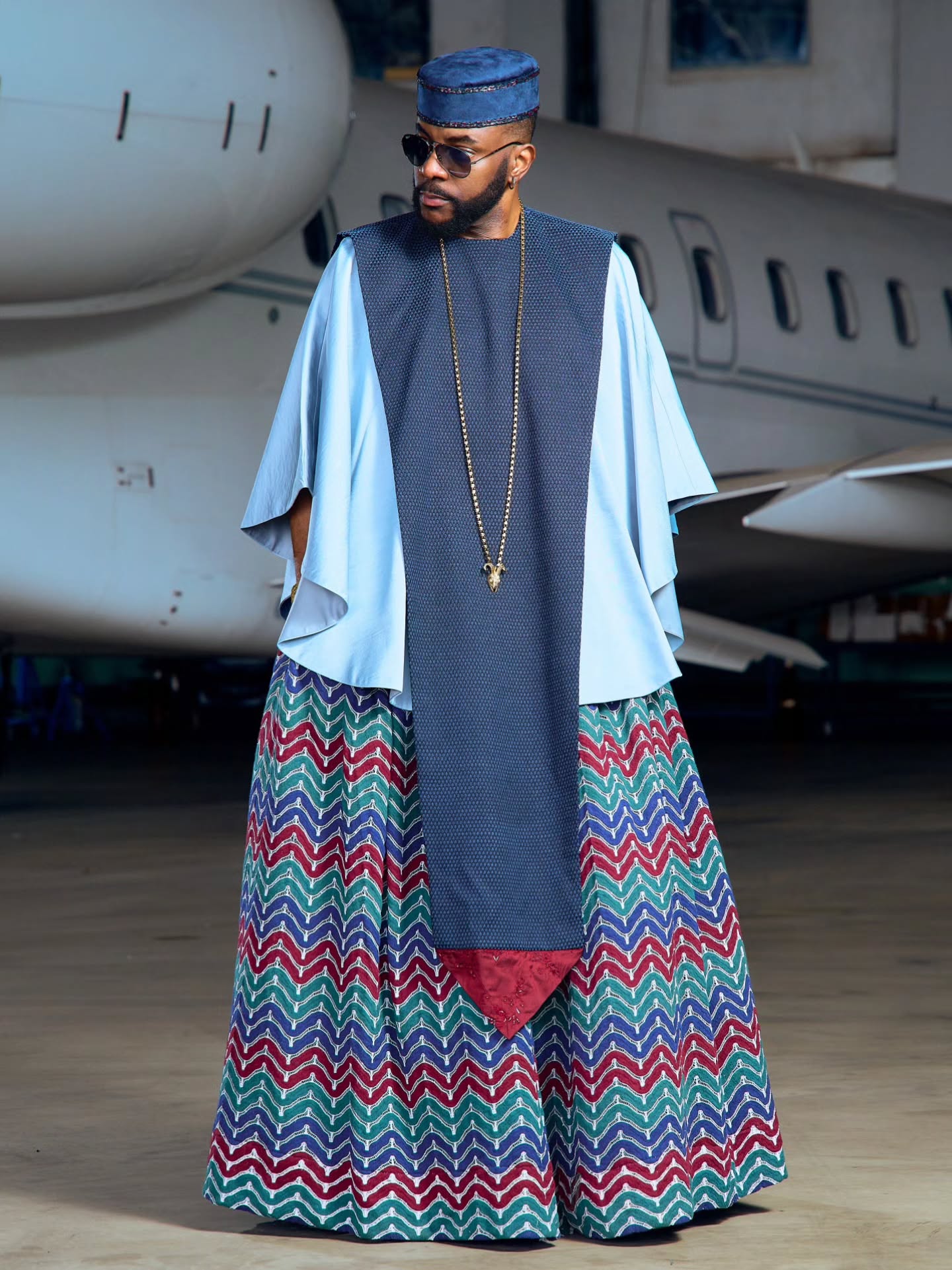 Ebuka Obi-Uchendu standing in a sky-blue Ugo Monye tunic, navy textured panel, and wide multi-coloured chevron trousers.