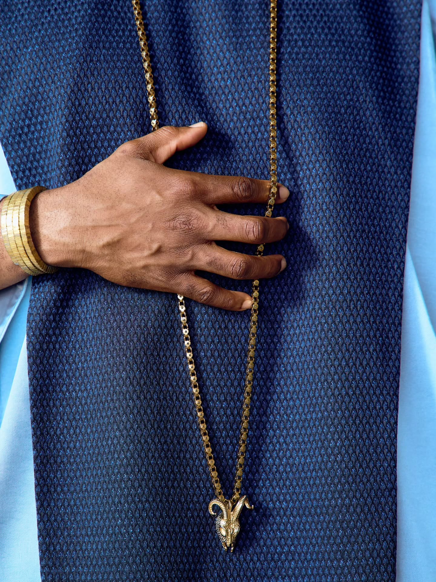 Long gold chain with a ram’s head pendant resting on a navy textured chest panel and a gold ribbed cuff on the wrist.