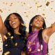 Two joyful African women cheering with arms raised amid falling gold confetti, celebrating two decades of BellaNaija.