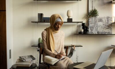 A Muslim woman in a hijab focused on studying in a modern indoor setting with a laptop and notebooks.