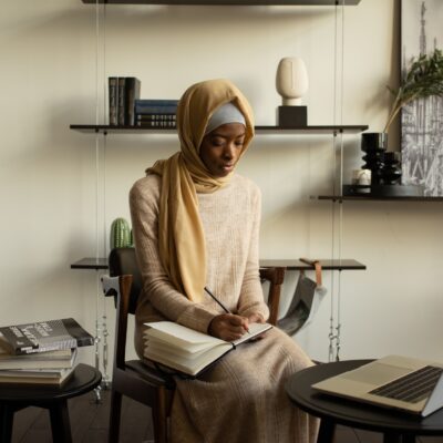 A Muslim woman in a hijab focused on studying in a modern indoor setting with a laptop and notebooks.